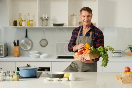 Foto de Sonriente hombre maduro sosteniendo cesta de verduras frescas al estar de pie en el mostrador de la cocina - Imagen libre de derechos