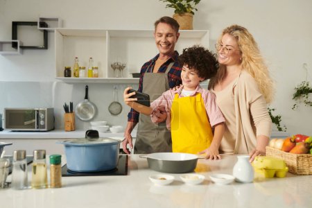 Photo for Cheerful preteen boy taking selfie with parents when they are cooking in kitchen - Royalty Free Image