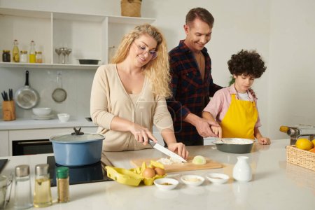 Téléchargez les photos : Femme souriante coupant l'oignon quand son mari et son fils mélangent farine et épices pour sauce bechamel - en image libre de droit