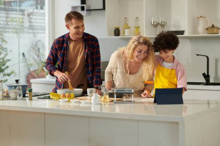 Foto de Familia alegre siguiendo la receta en la tableta digital al hacer pasta en casa - Imagen libre de derechos