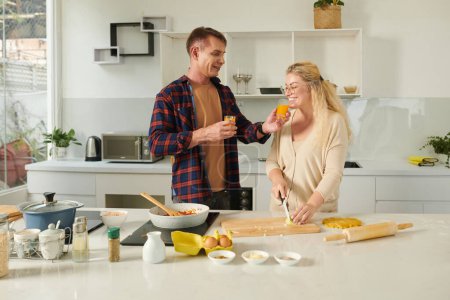 Foto de Cuidar hombre dando cocina esposa cortar cebollas taza de jugo de fruta fresca para oler para evitar llorar - Imagen libre de derechos