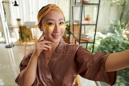 Téléchargez les photos : Jeune femme souriante en robe de soie prenant selfie après avoir appliqué des patchs sous les yeux - en image libre de droit