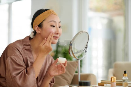 Téléchargez les photos : Sourire jeune femme regardant miroir grossissant lors de l'application de crème hydratante visage - en image libre de droit