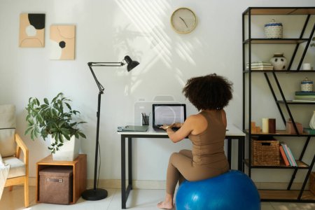 Téléchargez les photos : Femme assise sur un ballon de fitness lorsque vous travaillez sur un ordinateur portable à la maison, vue de derrière - en image libre de droit