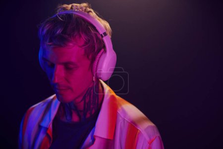 Téléchargez les photos : Portrait de jeune homme debout dans la lumière du néon et écoutant de la musique calme dans les écouteurs - en image libre de droit