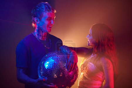 Foto de Feliz pareja joven con bola disco de pie en la pista de baile en el club nocturno y mirándose el uno al otro - Imagen libre de derechos