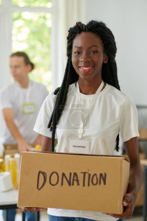 Téléchargez les photos : Portrait de heureuse jeune femme noire tenant boîte avec des produits donnés - en image libre de droit