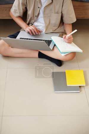 Photo for Cropped image of teenage boy sitting on floor in bedroom, watching webinar on laptop and taking notes in textbook - Royalty Free Image