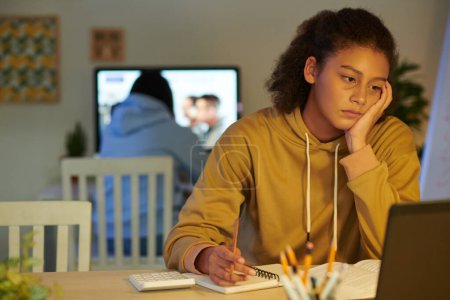 Téléchargez les photos : Fatigué et fatigué écolière regarder webinaire sur ordinateur portable et de prendre des notes dans le manuel - en image libre de droit