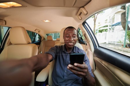 Foto de Sonriente hombre negro conduciendo coche y leyendo mensajes de texto en el teléfono inteligente - Imagen libre de derechos