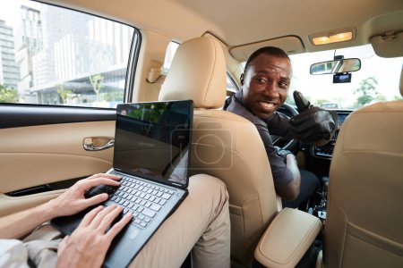 Téléchargez les photos : Chauffeur de taxi se retournant vers le passager travaillant sur un ordinateur portable et montrant pouces levés - en image libre de droit