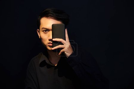 Téléchargez les photos : Sérieux jeune homme asiatique debout dans la chambre noire et couvrant un oeil avec smartphone - en image libre de droit