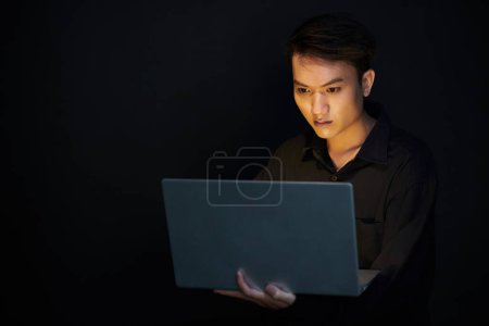 Téléchargez les photos : Sérieux jeune homme asiatique travaillant sur ordinateur portable dans la chambre noire - en image libre de droit