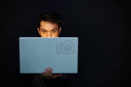 Téléchargez les photos : Pirate sérieux regardant la caméra sur l'écran d'ordinateur portable lorsque vous travaillez dans la pièce sombre - en image libre de droit