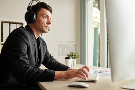 Photo for Concentrated businessman in headset looking at computer screen and typing - Royalty Free Image