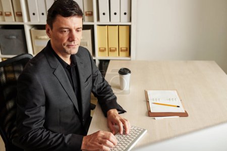 Téléchargez les photos : Homme d'affaires sérieux vérifiant ses e-mails sur ordinateur et prenant des notes dans le planificateur le matin - en image libre de droit