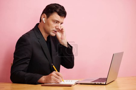 Téléchargez les photos : Homme d'affaires dans les écouteurs parler à un collègue, la lecture sur l'écran de l'ordinateur portable et de prendre des notes en plnner - en image libre de droit