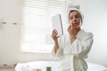 Foto de Mujer en albornoz con suave toalla blanca en la cabeza mirando en el espejo mientras está de pie delante de la cámara en el baño y tocando su cara - Imagen libre de derechos