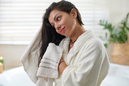 Foto de Joven mujer bonita en albornoz blanco secando su largo cabello oscuro con una toalla suave después de lavarlos mientras está de pie en el baño - Imagen libre de derechos