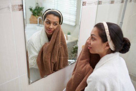 Téléchargez les photos : Brunette fille en peignoir toucher visage propre avec serviette douce après avoir lavé et regarder dans le miroir tout en se tenant dans la salle de bain - en image libre de droit