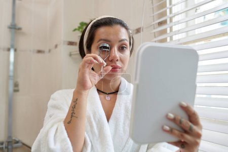 Téléchargez les photos : Jeune femme en peignoir blanc doux regardant dans le miroir dans sa main et en utilisant friseur de cils tout en se tenant dans la salle de bain - en image libre de droit