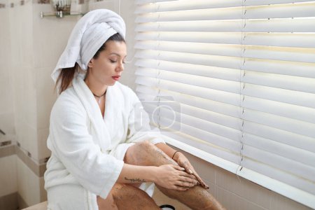 Foto de Mujer joven en albornoz sentada junto a la ventana con persianas venecianas en el baño y aplicando exfoliación corporal o descamación en las piernas - Imagen libre de derechos