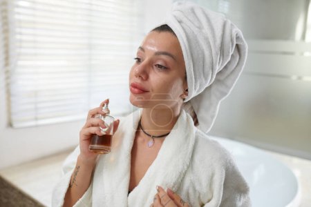 Foto de Joven mujer complacida en albornoz rociando nueva fragancia de la botella en su piel y disfrutando del olor del perfume mientras está de pie en el baño - Imagen libre de derechos