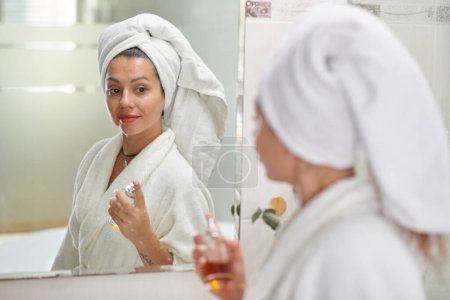 Foto de Mujer joven con toalla en la cabeza de pie delante del espejo en el baño y rociando la fragancia de la botella en su piel después de la ducha - Imagen libre de derechos