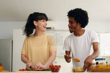 Foto de Dos felices citas interculturales jóvenes en camisetas mirándose entre sí mientras chica picando tomates cherry y hombre mezclando ingredientes de ensalada - Imagen libre de derechos