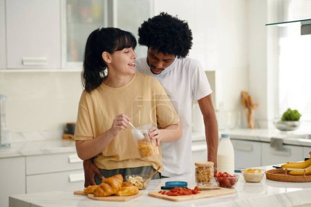 Téléchargez les photos : Jeune femme souriante regardant son mari afro-américain tout en restant à table dans la cuisine et en préparant le petit déjeuner - en image libre de droit
