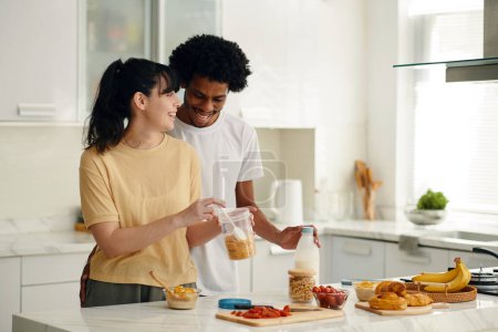 Téléchargez les photos : Joyeux jeune femme avec pot en plastique de flocons de maïs debout à côté de son mari et préparer le petit déjeuner par table de cuisine - en image libre de droit