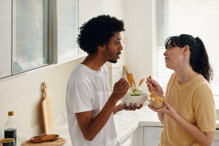 Photo for Side view of young multicultural couple with salad in bowls having breakfast and looking at one another in the kitchen - Royalty Free Image