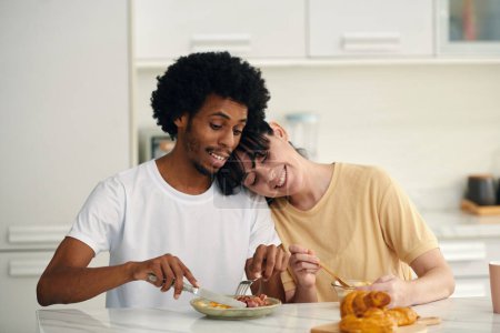 Photo for Young smiling woman keeping head on shoulder of her husband cutting small piece of homemade pizza for his sweetheart - Royalty Free Image