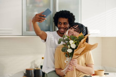 Téléchargez les photos : Jeune couple interculturel avec des fleurs blanches regardant l'écran du smartphone tout en se tenant dans la cuisine et faire du selfie - en image libre de droit
