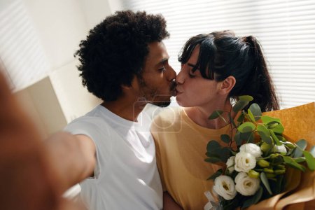 Téléchargez les photos : Jeune homme afro-américain et sa femme avec un bouquet de fleurs embrassant sur les lèvres tout en posant pour selfie devant la caméra à la maison - en image libre de droit