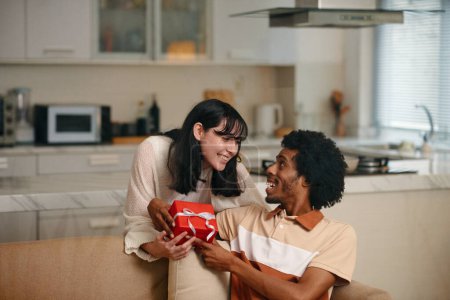 Photo for Young affectionate husband and wife looking at each other while African American man taking red giftbox being passed by happy woman - Royalty Free Image