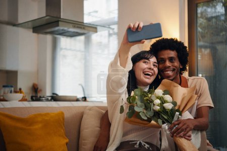 Téléchargez les photos : Femme heureuse avec bouquet de fleurs tenant smartphone devant elle et prenant selfie avec mari affectueux assis à côté d'elle - en image libre de droit