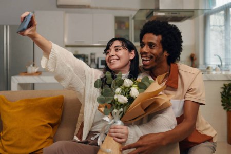 Foto de Joven mujer sonriente con flores blancas tomando selfie con su marido en casa mientras sostiene el teléfono inteligente frente a sí misma - Imagen libre de derechos