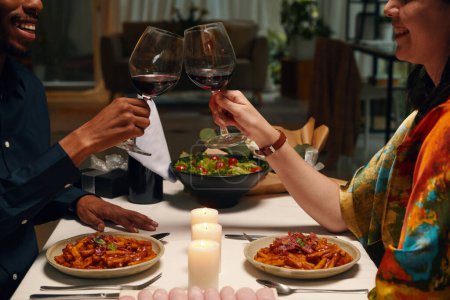 Photo for Cropped shot of happy young intercultural valentines clinking with glasses of red wine while sitting by served table with homemade food - Royalty Free Image