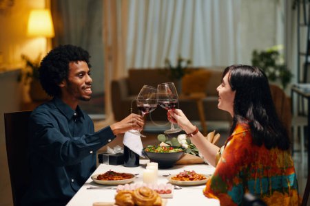 Foto de Vista lateral de los felices San Valentín interculturales jóvenes mirándose unos a otros y tintineando con copas de vino tinto sobre la mesa festiva servida - Imagen libre de derechos