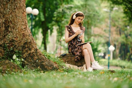 Téléchargez les photos : Jeune femme souriante en tenue décontractée et écouteurs assis sous un arbre dans le parc, écoutant de la musique et lisant un livre de contes ou un journal - en image libre de droit