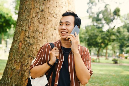 Téléchargez les photos : Souriant asiatique gars avec sac à dos debout contre tronc d'arbre dans le parc et parler sur smartphone avec un ami tout en regardant de côté - en image libre de droit