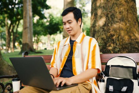 Téléchargez les photos : Jeune homme d'affaires asiatique en tenue décontractée regardant écran d'ordinateur portable et tapant tout en étant assis sur le banc dans le parc avec de nombreux arbres - en image libre de droit