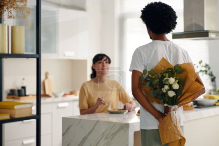 Téléchargez les photos : Dos du jeune Afro-Américain tenant un bouquet de roses blanches pour sa femme ou sa petite amie assise à table devant lui - en image libre de droit