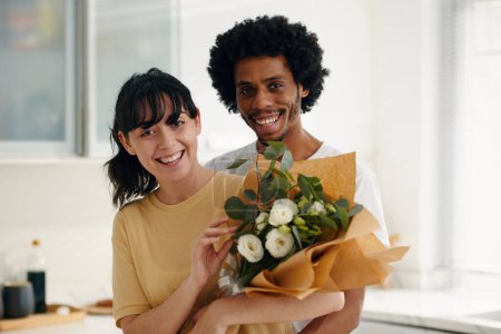 Foto de Joven y alegre esposo y esposa intercultural con ramo de flores blancas envueltos en papel mirando a la cámara mientras están de pie en la cocina - Imagen libre de derechos