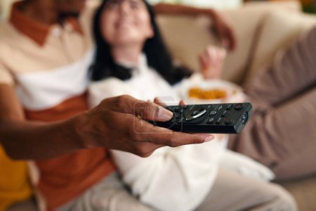 Téléchargez les photos : Main de jeune homme afro-américain avec télécommande choisir canal tout en étant assis à côté de sa femme devant le téléviseur à la maison - en image libre de droit