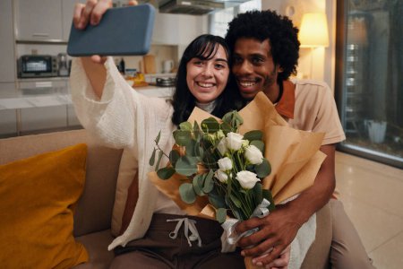 Photo for Happy young woman with bouquet of white flowers holding smartphone in front of herself while taking selfie with affectionate husband - Royalty Free Image