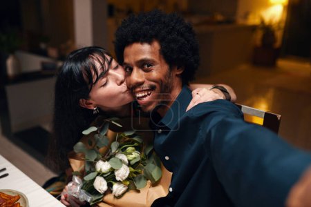 Foto de Joven hombre afroamericano sonriente mirando a la cámara mientras toma selfie con su esposa cariñosa con flores besándolo en la mejilla - Imagen libre de derechos