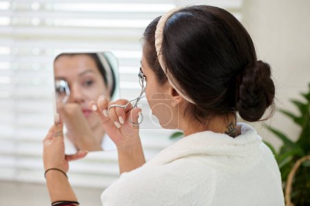 Téléchargez les photos : Brunette fille en peignoir blanc friser les cils et regarder dans le miroir dans sa main tout en prenant soin de sa beauté dans la salle de bain - en image libre de droit