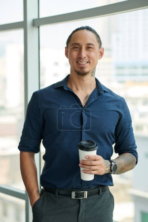 Téléchargez les photos : Jeune employé masculin ou solopreneur en chemise bleu foncé regardant la caméra avec le sourire à la pause café tout en se tenant contre une grande fenêtre - en image libre de droit
